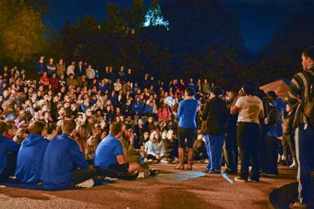 Students crowd around the clock tower at night while a speaker stands at a microphone.